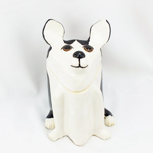 May include: A ceramic figurine of a dog with black and white fur. The dog is sitting with its legs tucked under its body and has a white collar. The dog has large, brown eyes and a black nose.
