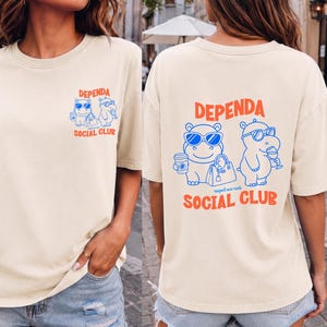 May include: A cream-colored t-shirt featuring a blue and orange graphic of two hippos wearing sunglasses. The front chest displays a small version of the design, while the back features a larger version with the text "DEPENDA SOCIAL CLUB".