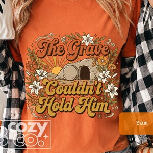 May include: An orange t-shirt featuring a graphic print of a stone tomb with a sunburst, surrounded by colorful flowers and greenery. The text reads, The Grave Couldn't Hold Him, in a retro font. A black and white plaid shirt is worn over the t-shirt.