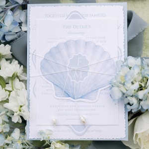 May include: A rectangular wedding invitation card with a light blue scalloped border, featuring a central white card with a large blue seashell illustration. The seashell displays the initials FM. A thin white string with two small pearls ties the cards.