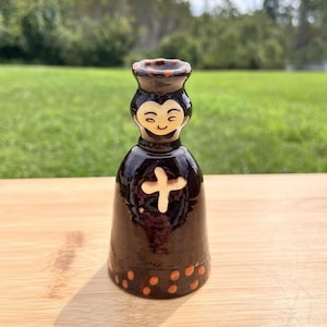 May include: A dark brown glazed ceramic figurine shaped like a person with a bell-like body. The figure features a cream-colored cross on the chest, a smiling face, and a rim decorated with small orange dots at the top and bottom edges.