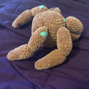 May include: A brown plush toy with a soft, textured surface and multiple limbs. The stuffed creature features small, light teal accents on the tips of its appendages and joints, providing a subtle contrast to the solid brown color of the body.