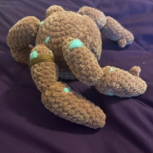 May include: A brown crocheted plush toy with a round body and multiple segmented limbs. The plush features small, light blue embroidered accents scattered across its surface and a single dark brown yarn band wrapped around one of its limbs.