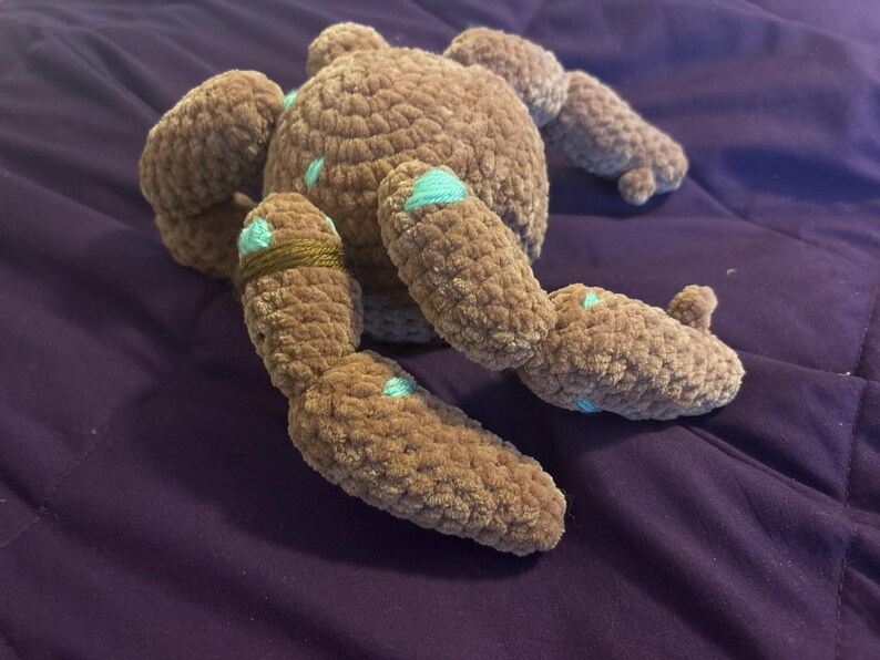 May include: A brown crocheted plush toy with a round body and multiple segmented limbs. The plush features small, light blue embroidered accents scattered across its surface and a single dark brown yarn band wrapped around one of its limbs.