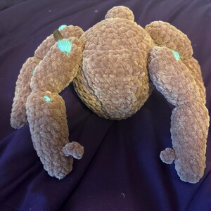 May include: A plush spider toy crafted from brown, textured yarn. The creature features a rounded central body and eight segmented legs extending outward. Small, bright turquoise accents are visible at the joints of several legs.