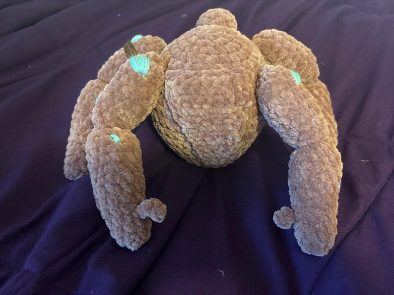 May include: A plush spider toy crafted from brown, textured yarn. The creature features a rounded central body and eight segmented legs extending outward. Small, bright turquoise accents are visible at the joints of several legs.