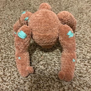 May include: A plush toy shaped like a spider, crafted from soft, brown, velvet-like yarn. The creature features a rounded central body with eight extended legs, accented with small, embroidered patches of bright turquoise thread throughout.