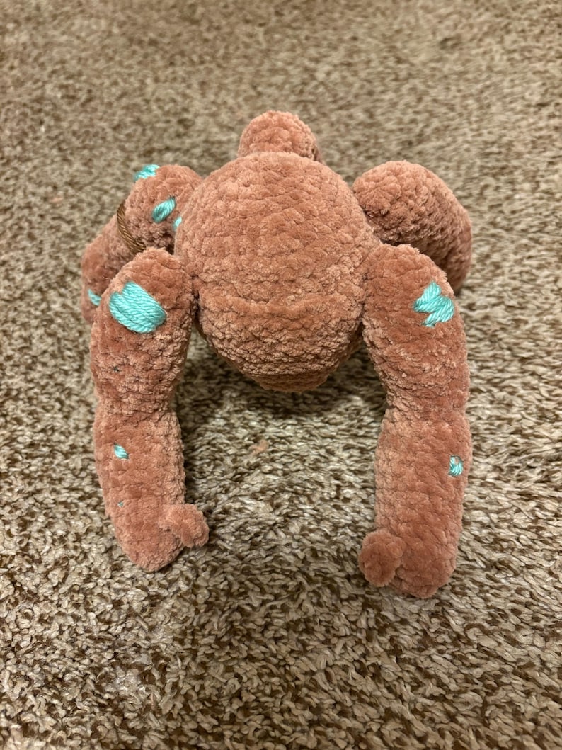 May include: A plush toy shaped like a spider, crafted from soft, brown, velvet-like yarn. The creature features a rounded central body with eight extended legs, accented with small, embroidered patches of bright turquoise thread throughout.