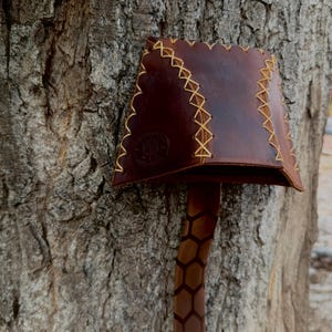 May include: A dark brown leather object with a flared top section featuring light-colored cross-stitching along the edges and a circular embossed logo. A long, narrow strap extends downward, decorated with a repeating dark brown hexagonal pattern.