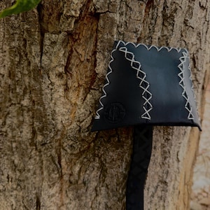 May include: A black leather pouch with a trapezoidal shape, featuring light-colored cross-stitching along the edges and a central seam. A long, narrow black strap with a repeating diamond pattern extends downward from the bottom of the pouch.
