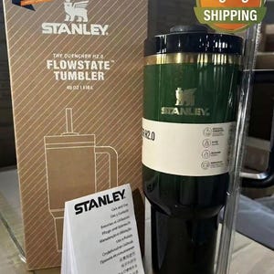 May include: A dark green Stanley The Quencher H2.0 FlowState tumbler with a black lid and handle, accompanied by its cardboard packaging box, a clear plastic straw, and a white care and use instruction booklet with the Stanley brand logo.