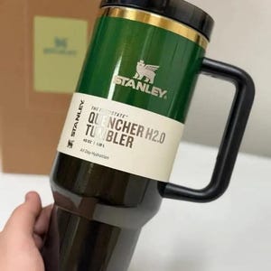 May include: A Stanley Quencher H2.0 tumbler with a dark green upper section and a black lower section, featuring a black handle and lid. A white product label displays the text Stanley, The Quencher H2.0 Tumbler, 40 oz, 1.18 L, All Day Hydration.