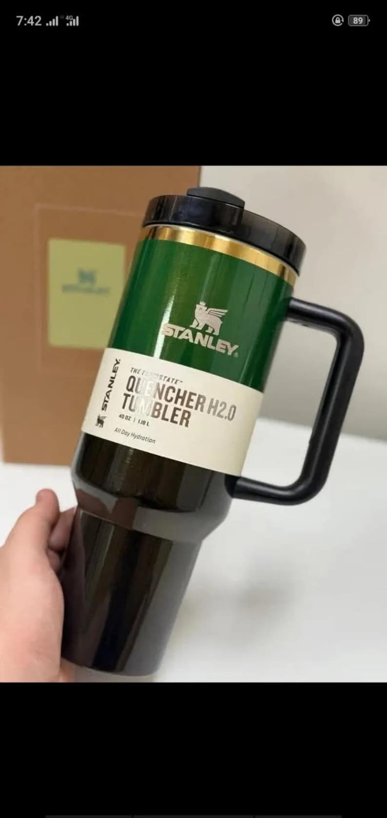 May include: A Stanley Quencher H2.0 tumbler with a dark green upper section and a black lower section, featuring a black handle and lid. A white product label displays the text Stanley, The Quencher H2.0 Tumbler, 40 oz, 1.18 L, All Day Hydration.
