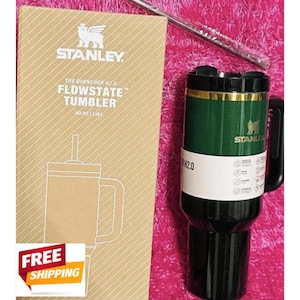 May include: A dark green Stanley Quencher H2.0 FlowState tumbler with a black handle, lid, and base, featuring a gold rim and logo. It is accompanied by its tan cardboard packaging box labeled 40 oz and a clear straw in a plastic sleeve.