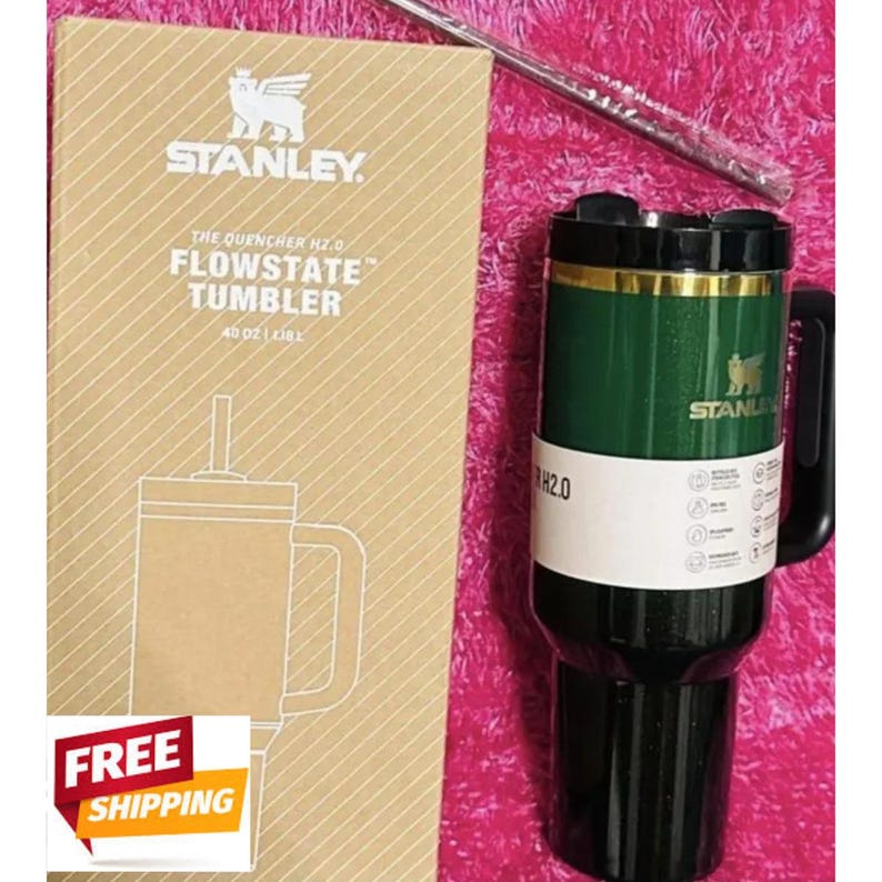 May include: A dark green Stanley Quencher H2.0 FlowState tumbler with a black handle, lid, and base, featuring a gold rim and logo. It is accompanied by its tan cardboard packaging box labeled 40 oz and a clear straw in a plastic sleeve.