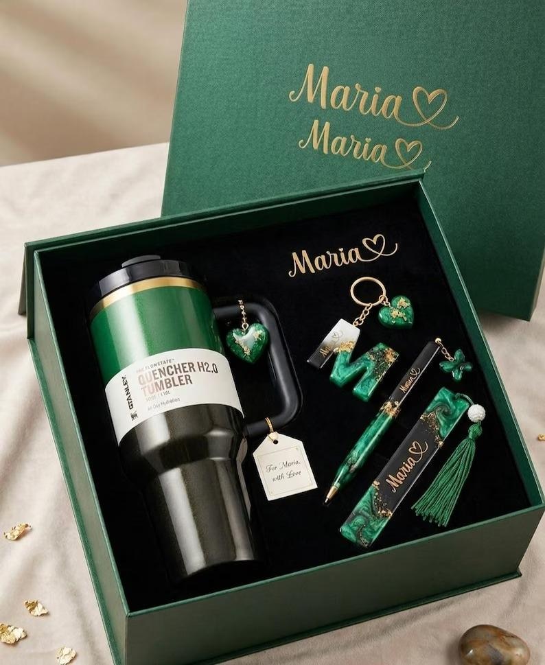 May include: A dark green gift box containing a Stanley Quencher H2.0 tumbler, a letter M keychain, a pen, and a bookmark. The items feature green and black marbled resin with gold accents. The name Maria is printed on the box and items.