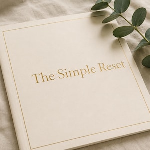 May include: A rectangular book with a cream-colored cover featuring the gold-embossed text The Simple Reset centered on the front. The cover is framed by a thin gold border line. A green eucalyptus branch with small oval leaves rests nearby.