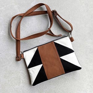Può includere: Una borsa a tracolla geometrica con un cinturino in pelle marrone. La borsa presenta un design a blocchi di colore nero, bianco e marrone. La borsa ha una chiusura a zip e un cinturino in pelle marrone.
