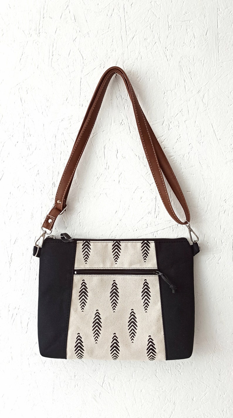 May include: A black and white patterned crossbody bag with a brown leather strap. The bag has a zipper closure and two pockets on the front.