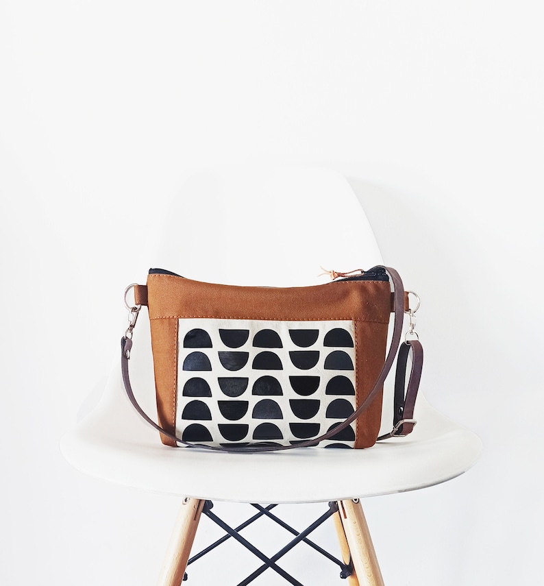 May include: A brown canvas crossbody bag with a black and white geometric pattern. The bag has a long adjustable strap and a zipper closure.