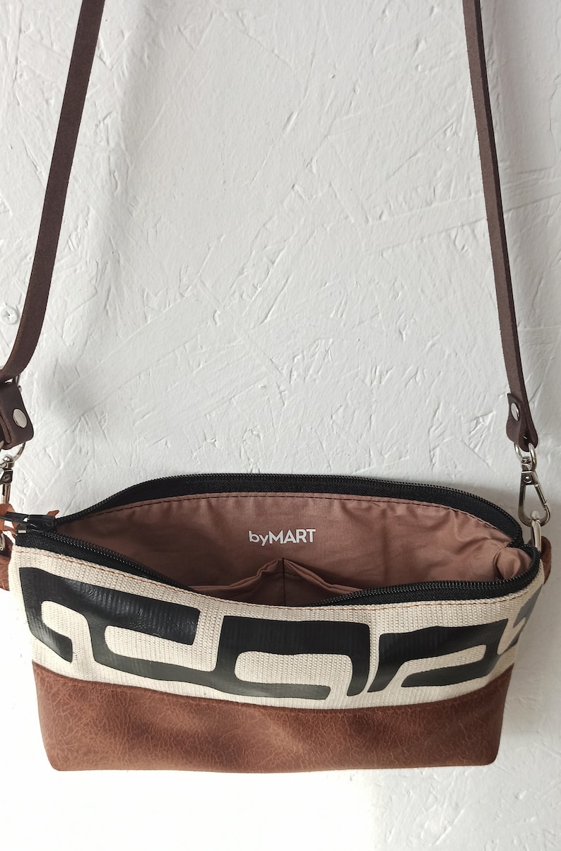 Pode incluir: Bolsa transversal aberta com um padr&atilde;o geom&eacute;trico em preto e branco. A bolsa tem uma base e uma al&ccedil;a de couro marrom. O interior &eacute; marrom com o texto "byMART".