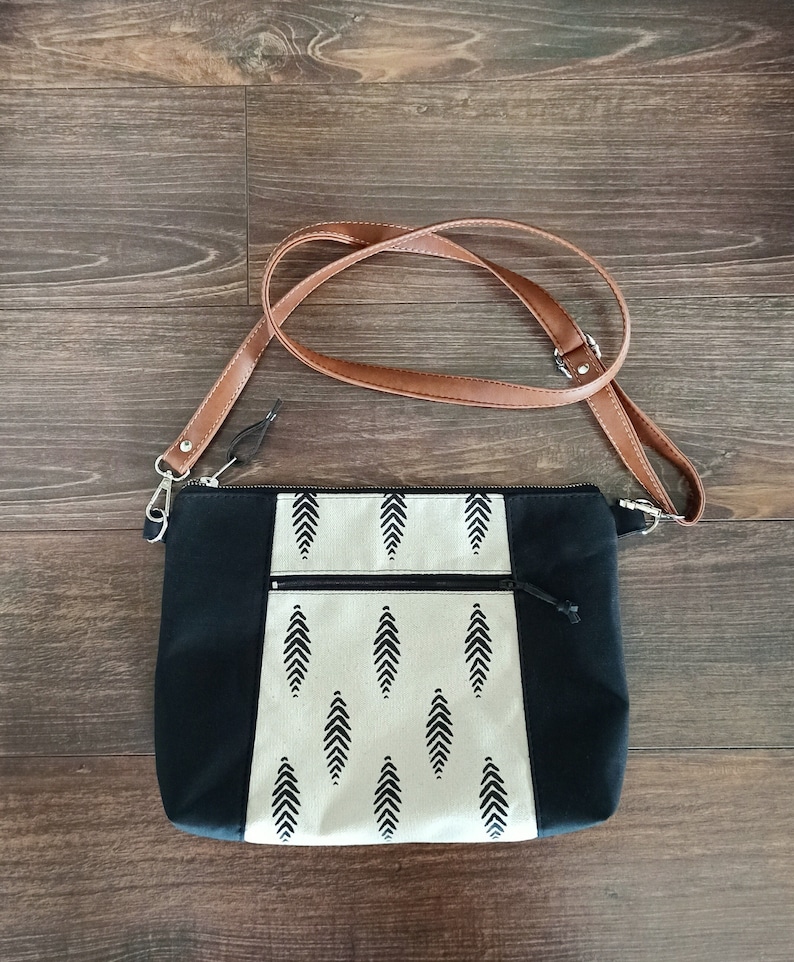 May include: A black and white patterned crossbody bag with a brown leather strap. The bag has a zipper closure and a small pocket on the front.
