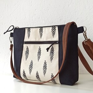 May include: A black and white canvas tote bag with a brown leather strap. The bag has a zipper closure and a small pocket on the front. The canvas features a black and white geometric pattern.