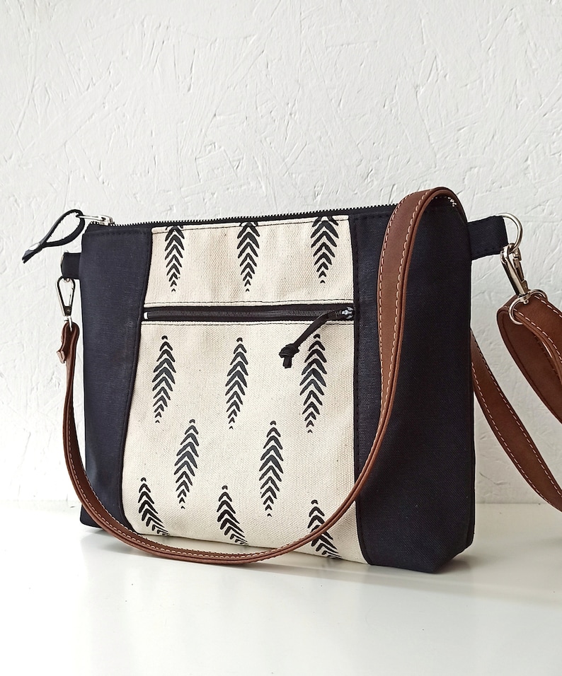 May include: A black and white canvas tote bag with a brown leather strap. The bag has a zipper closure and a small pocket on the front. The canvas features a black and white geometric pattern.