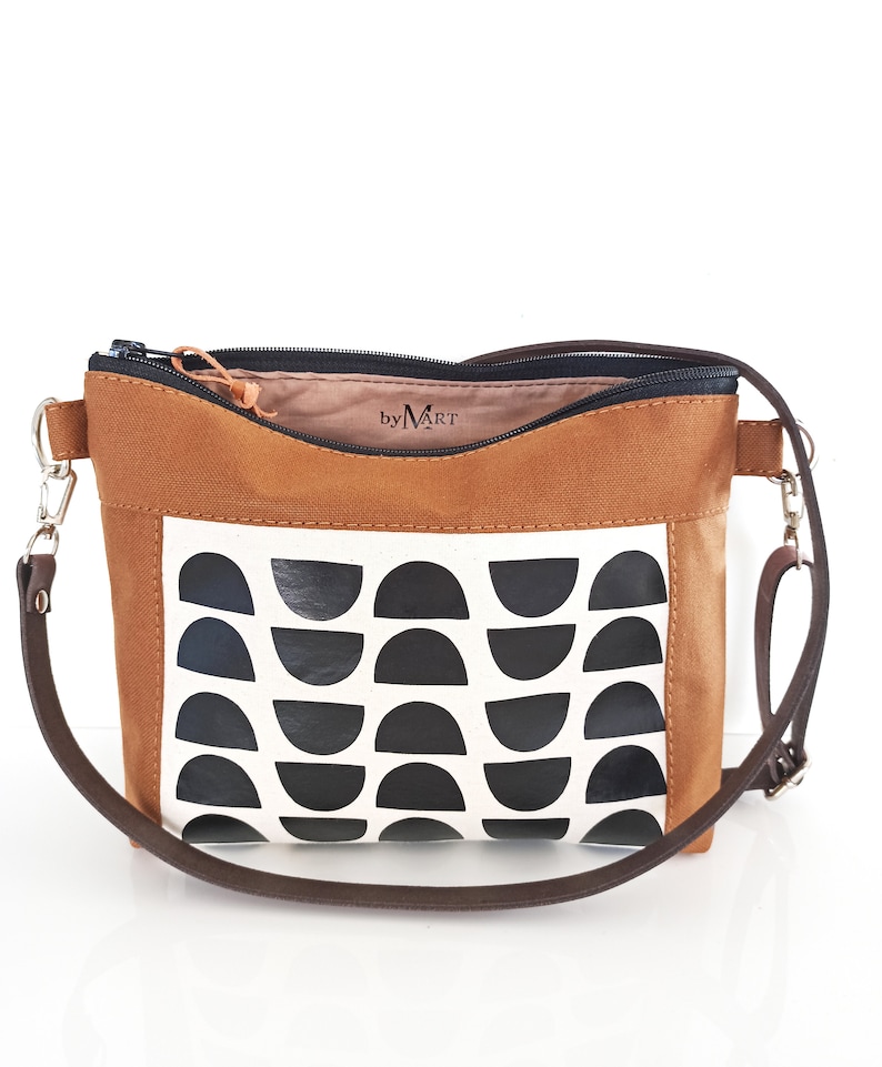 May include: A brown canvas crossbody bag with a black and white geometric pattern. The bag has a zipper closure and a brown adjustable strap. The bag has a label that says "by MART".
