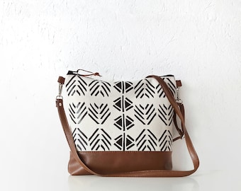 May include: A crossbody bag featuring a white canvas body with a black geometric chevron and diamond pattern. The base and the adjustable shoulder strap are made of brown material. The bag includes a top zipper closure with a small brown pull tab.