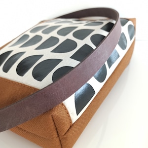May include: A brown canvas tote bag with a black and white geometric pattern. The bag has a brown leather strap.