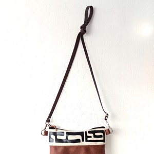 Pode incluir: Uma bolsa transversal marrom e preta com um design geom&eacute;trico. A bolsa apresenta uma base marrom tipo camur&ccedil;a, uma se&ccedil;&atilde;o central creme com formas pretas e uma al&ccedil;a marrom escura. A bolsa est&aacute; pendurada contra uma parede branca.