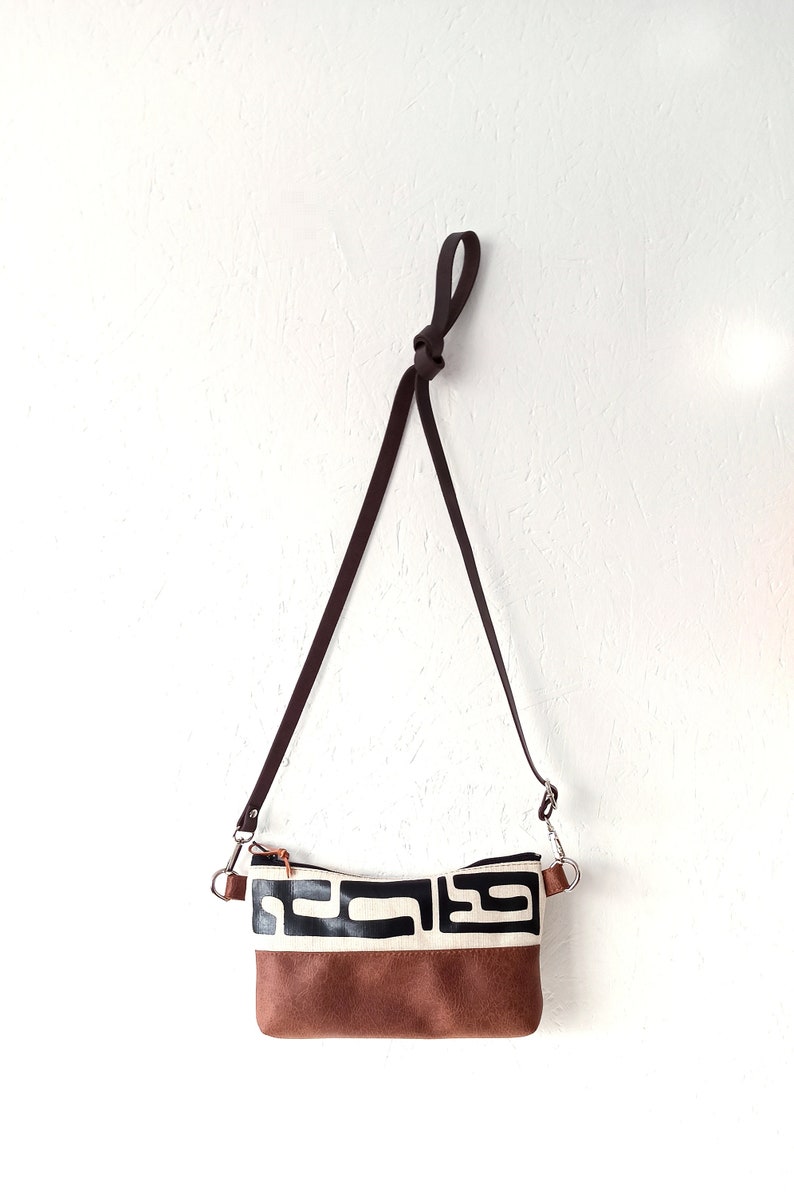 Pode incluir: Uma bolsa transversal marrom e preta com um design geom&eacute;trico. A bolsa apresenta uma base marrom tipo camur&ccedil;a, uma se&ccedil;&atilde;o central creme com formas pretas e uma al&ccedil;a marrom escura. A bolsa est&aacute; pendurada contra uma parede branca.