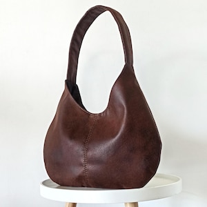 May include: A brown leather hobo bag with a single shoulder strap. The bag has a curved shape and a smooth texture. The bag is sitting on a white table with wooden legs. The bag is a medium size.