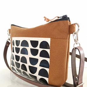 May include: A brown canvas crossbody bag with a black and white geometric pattern. The bag has a zipper closure and a brown leather strap.