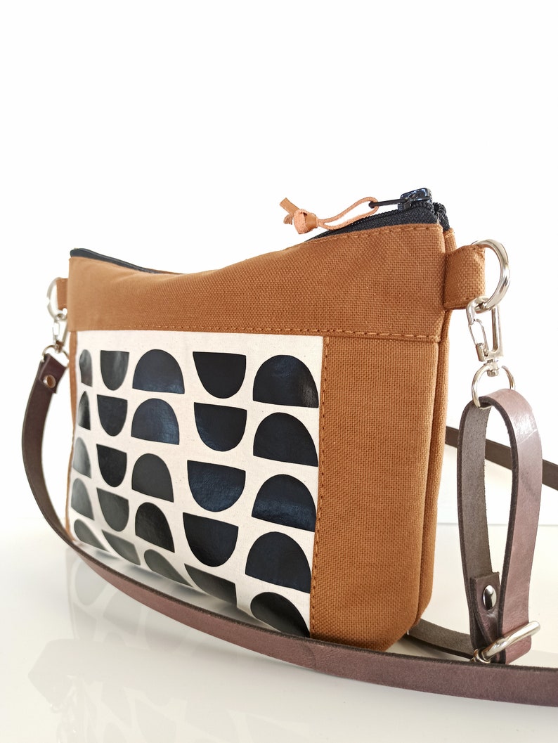 May include: A brown canvas crossbody bag with a black and white geometric pattern. The bag has a zipper closure and a brown leather strap.
