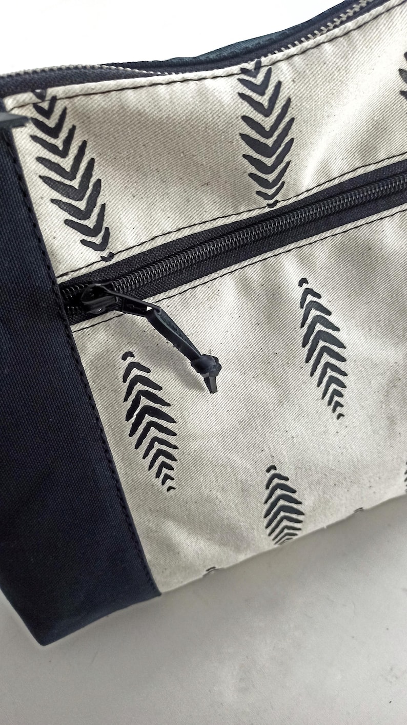 May include: A white canvas zippered pouch with a black zipper and black leaf-shaped patterns. The pouch has a black fabric border.
