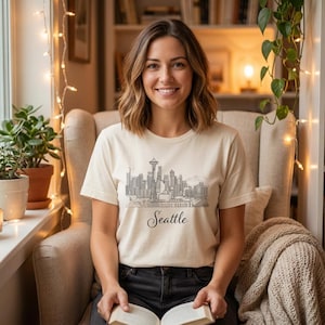 Könnte beinhalten: Ein cremefarbenes Kurzarm-T-Shirt mit einer schwarzen Strichzeichnung der Skyline von Seattle, einschließlich der Space Needle, und dem Wort Seattle in kursiver Schrift direkt unter den Gebäuden der Stadt.