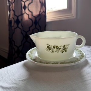 Peut inclure: Une saucière en verre opalin blanc avec sa soucoupe assortie, ornée d'un motif répétitif de petites fleurs vert olive. La saucière est dotée d'une anse unique et d'un bec verseur, tandis que la soucoupe présente une bordure florale.