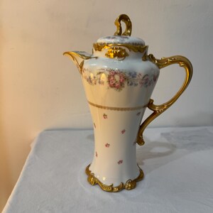 Puede incluir: Una cafetera de porcelana blanca con un delicado patrón floral de rosas rosadas y flores azules. La pieza presenta adornos dorados en el asa, el pico, la base y la tapa, la cual está rematada con un remate decorativo de color dorado.
