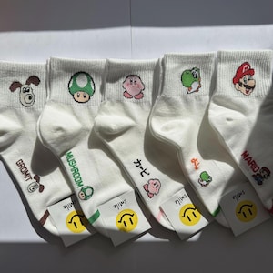 Puede incluir: Cinco pares de calcetines tobilleros blancos con bordados de arte en píxeles de personajes e iconos como Gromit, un champiñón verde, Kirby, Yoshi y Mario. Cada calcetín muestra el nombre del personaje en texto bajo el diseño.