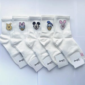 May include: Five pairs of white ankle socks, each featuring a small embroidered character face: Winnie the Pooh, Daisy Duck, Mickey Mouse, Donald Duck, and Piglet. Each sock has a white paper tag attached with the Disney logo printed on it.