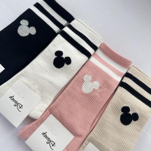 May include: Four pairs of crew socks featuring a ribbed texture and two stripes at the top. The colors include black, white, pink, and beige. Each sock displays a small embroidered Mickey Mouse head icon. Two pairs have white paper tags with the Disney logo.