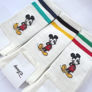 May include: Three pairs of white crew socks featuring an embroidered Mickey Mouse character wearing red shorts and yellow shoes. Each pair has a distinct striped cuff design in teal and navy, yellow and black, or green and red. A Disney tag is attached.
