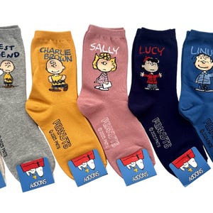 Peut inclure: Un ensemble de sept chaussettes montantes de différentes couleurs, dont beige, gris, jaune, rose, bleu marine, bleu et blanc. Chaque chaussette présente un personnage Peanuts, dont Charlie Brown, Sally, Lucy, Linus et Snoopy, avec leurs noms.