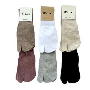 Pode incluir: Seis pares de meias com separação para os dedos em castanho, malva, branco, cinzento, bege e preto. Cada par está preso a um cartão com a etiqueta K'sox e Made in Korea. As meias apresentam uma separação entre o dedo grande e os outros.