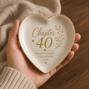 May include: A heart-shaped ceramic trinket dish in cream with a gold rim. The center features gold text reading "Chapter 40" above the phrase "Not just a number, a beautiful life so far," accented by a small gold leaf sprig and tiny heart icons.