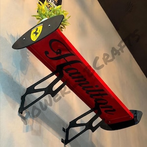 May include: A red automotive-style rear spoiler shelf featuring black support brackets and end plates. The top surface displays a yellow and black Ferrari logo, while the side panel features the black cursive text Hamilton.
