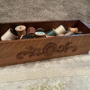 May include: A rectangular wooden box with a carved floral motif on the front panel, containing a collection of assorted sewing thread spools in various colors including white, green, black, and tan, resting inside the open storage container.