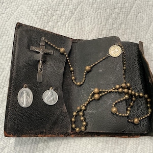 May include: A dark brown leather folding case containing a metal rosary with round beads, a crucifix, and two oval religious medals. The rosary features a circular medallion, while the crucifix is secured within a small leather strap inside.